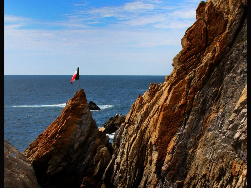 Cliff diver mid-dive from a rocky outcrop above the ocean in Acapulco, with a Mexican flag on a nearby rock