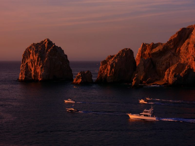 Fishing boats passing beneath dramatic rock formations at Land's End, Cabo San Lucas, glowing orange in the sunset light