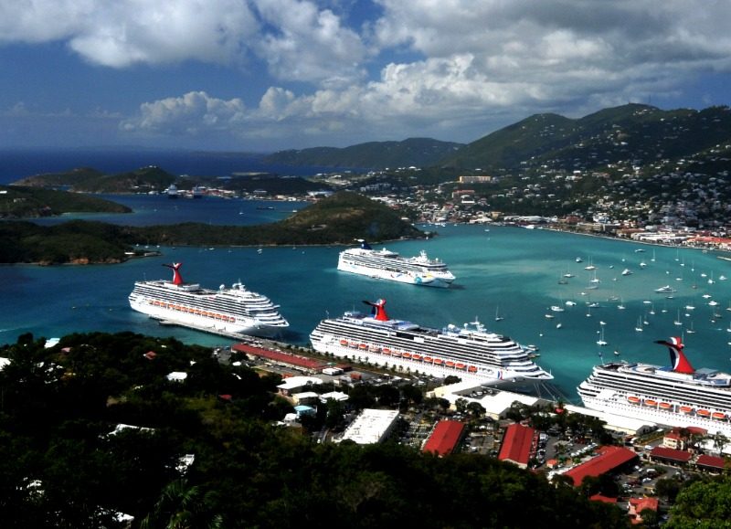 Aerial view of three cruise ships docked in a turquoise Caribbean harbor, surrounded by green hills and a hillside town