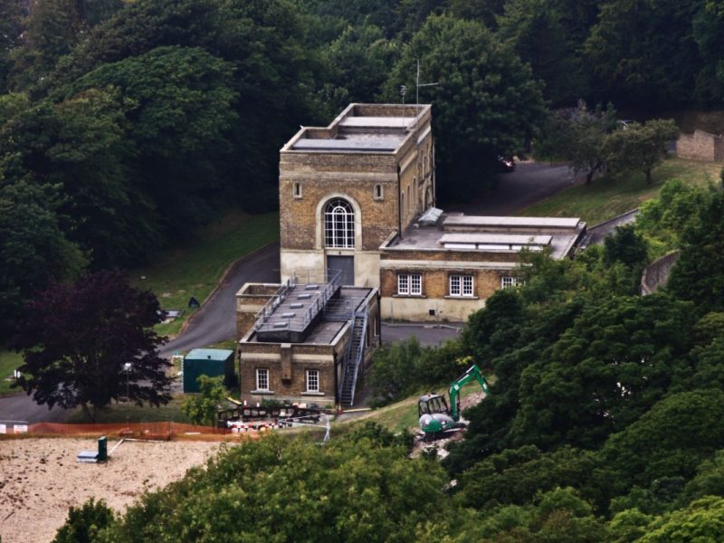 Brick building with a construction excavator beside it, set on a wooded hillside — no coast or mansion visible in frame