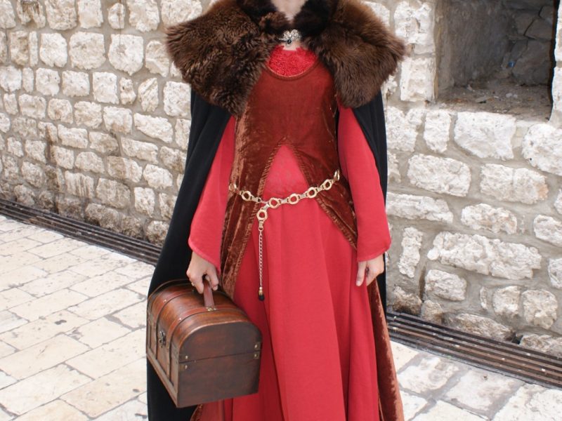 Woman posing in a medieval-style red velvet gown and fur-trimmed headpiece against a stone wall in Dubrovnik