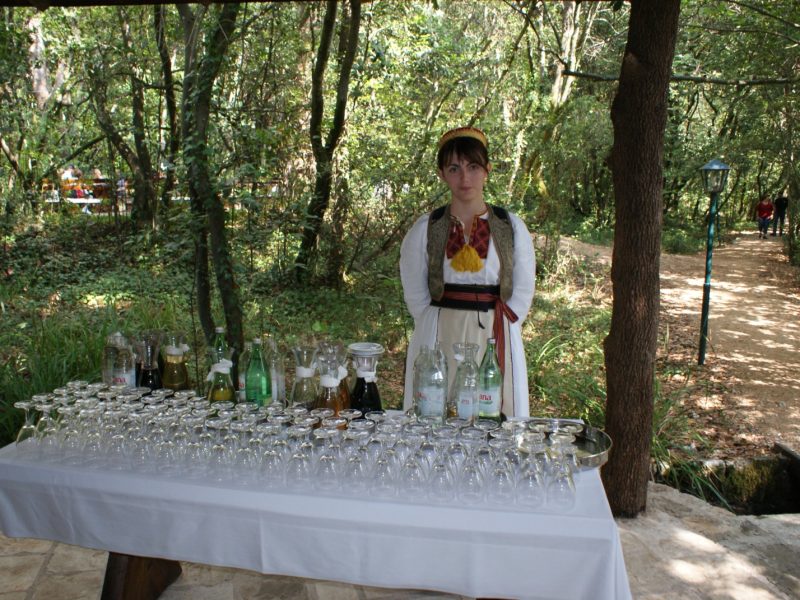 Woman in traditional dress standing behind a table set with wine glasses in a wooded outdoor setting