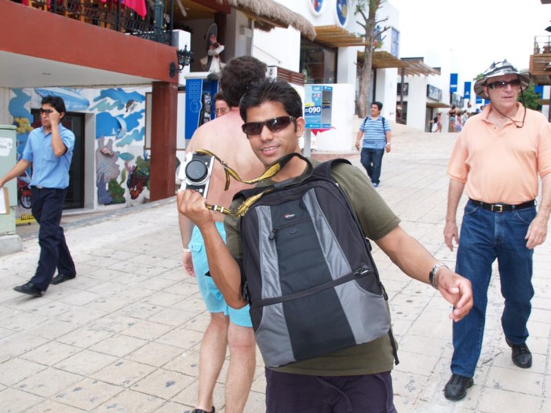 Tourist posing with a camera on a pedestrian street in a Mexican beach town, with shops and a thatched-roof restaurant behind him