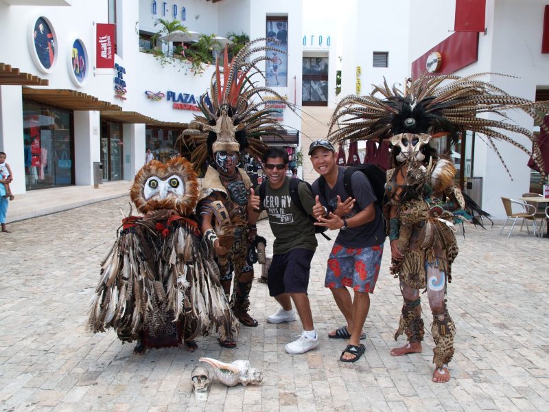 Tourists posing with performers in elaborate feathered Aztec-style headdresses and animal costumes in a Mexican plaza