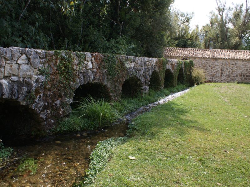 Old stone arch bridge crossing a narrow stream, surrounded by green lawns and trees
