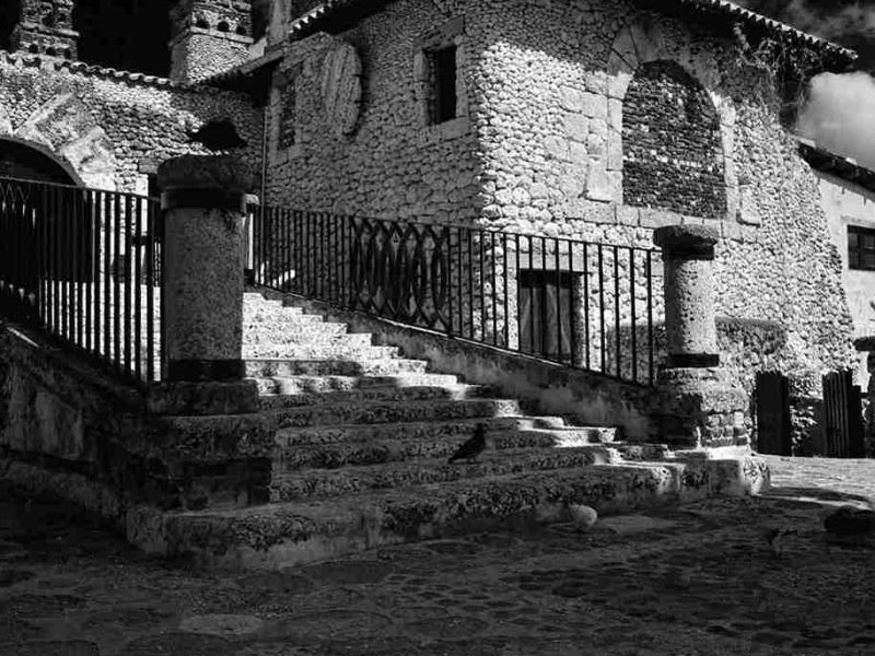 Black and white photo of a rustic stone building with a staircase and cobblestone courtyard