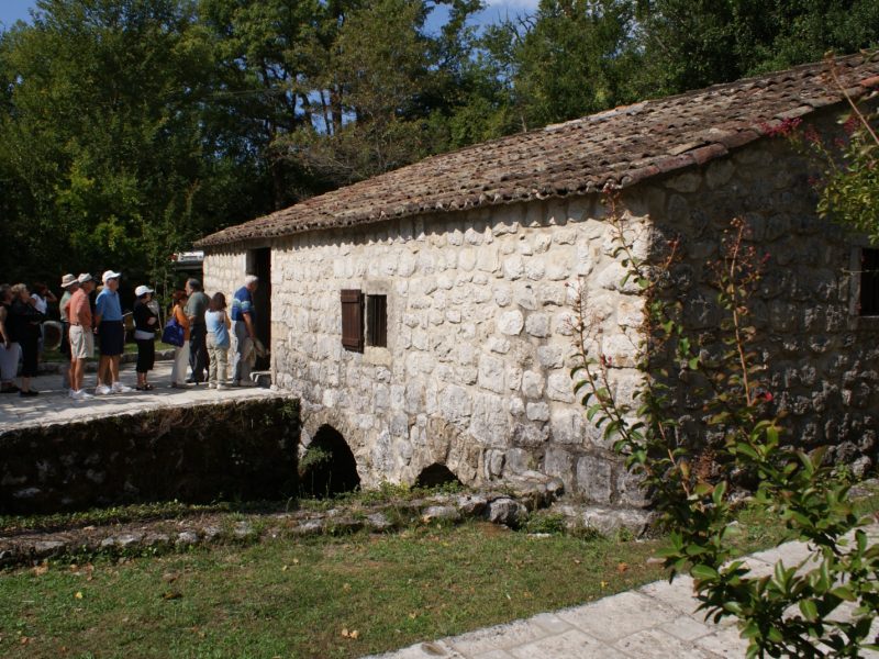 Group of tourists gathered outside a rustic stone watermill in a wooded setting
