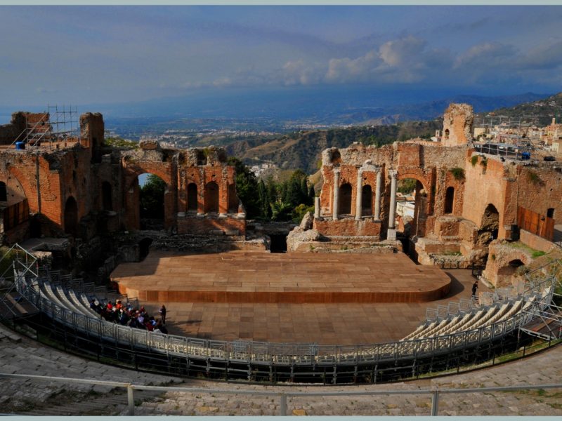 Ancient Greek-Roman theater in Taormina, Sicily, with brick ruins and stone columns framing a view of the coastline and mountains below
