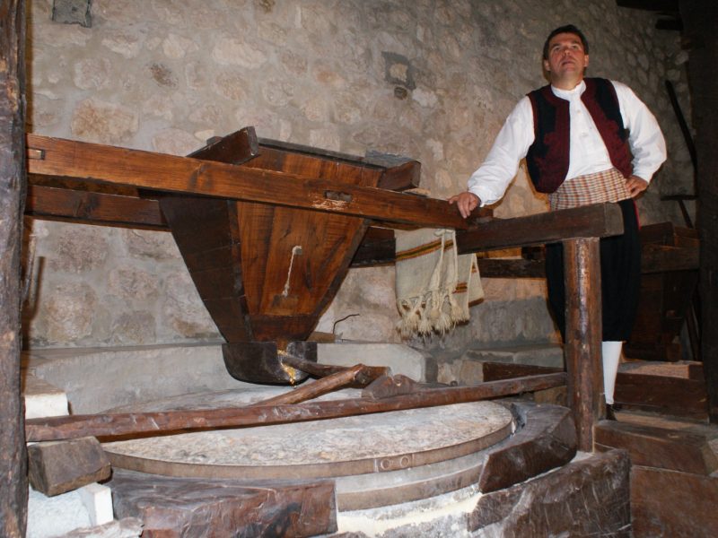 Man in traditional vest and shirt standing beside an old wooden olive or wine press