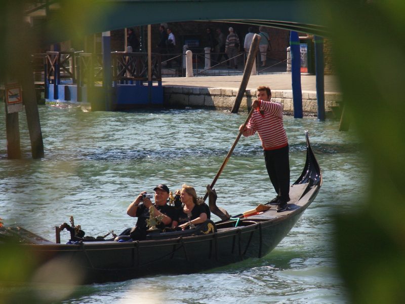 Gondolier in a striped shirt rowing two tourists past a bridge and waterfront promen