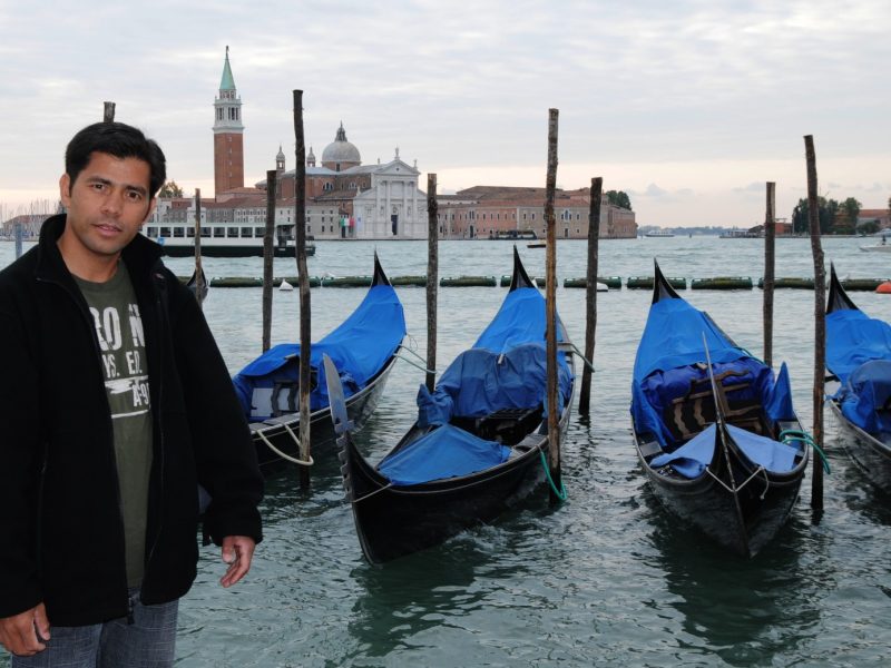Man posing beside moored gondolas in the Venice lagoon, with the Church of San Giorgio Maggiore visible across the water